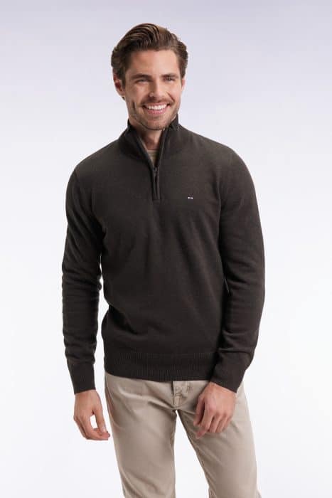 A man with short brown hair and a beard is smiling and wearing a dark quarter-zip sweater and beige pants, standing against a plain light background.