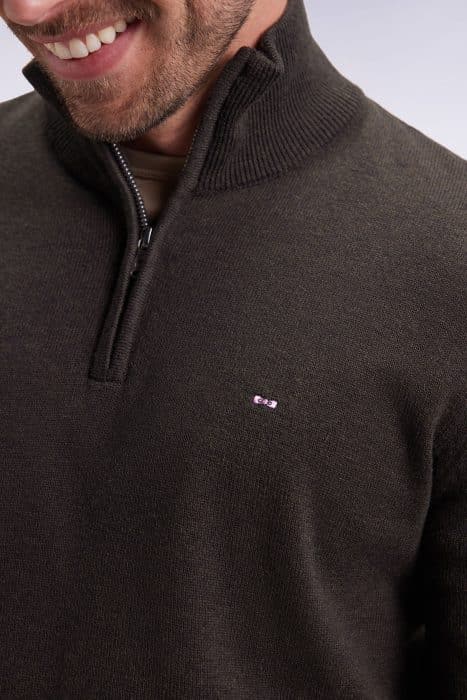 A close-up of a smiling man wearing a dark olive green quarter-zip sweater with a ribbed collar and a small embroidered logo on the chest.