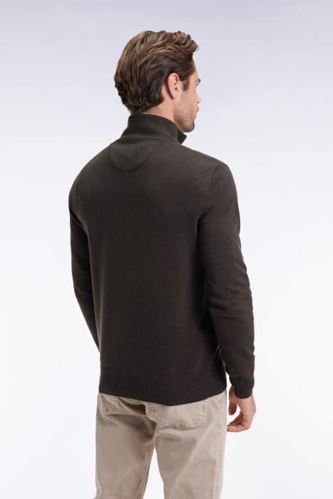 A man with short brown hair is standing with his back to the camera, wearing a dark long-sleeve sweater and light-colored pants against a plain, light background.