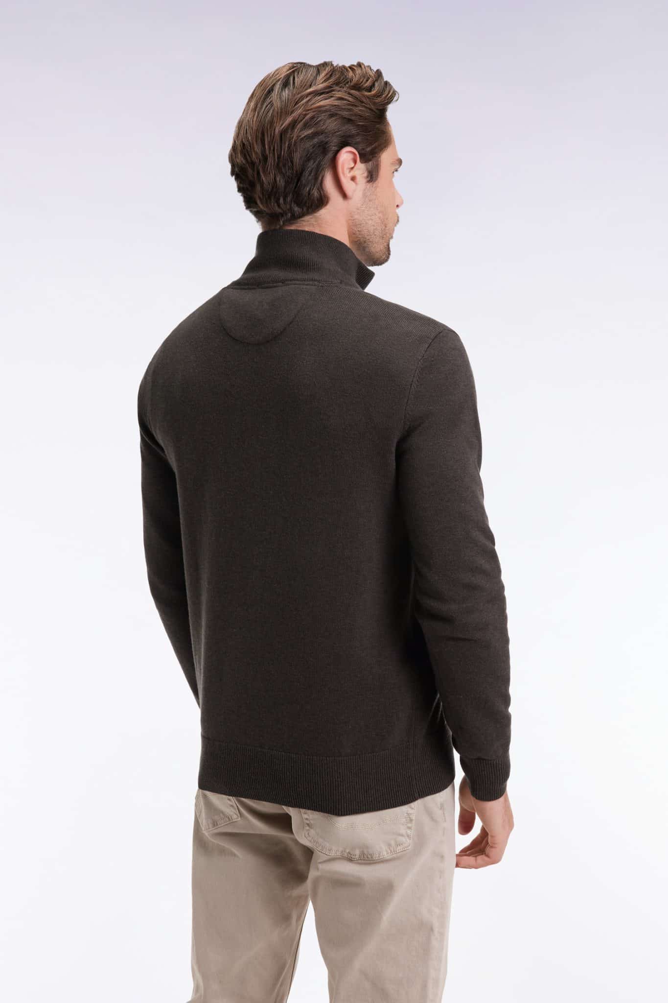 A man with short brown hair is standing with his back to the camera, wearing a dark long-sleeve sweater and light-colored pants against a plain, light background.