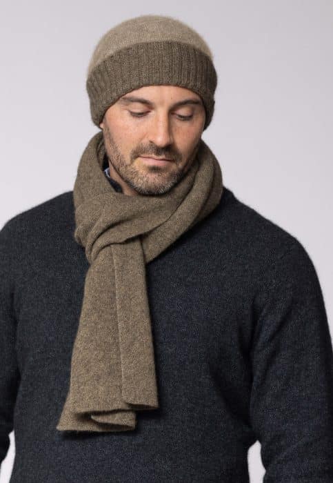 A man wearing a brown knit beanie and matching scarf over a dark sweater looks down with a neutral expression against a plain light background.