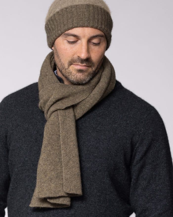 A man wearing a brown knit beanie and matching scarf over a dark sweater looks down with a neutral expression against a plain light background.