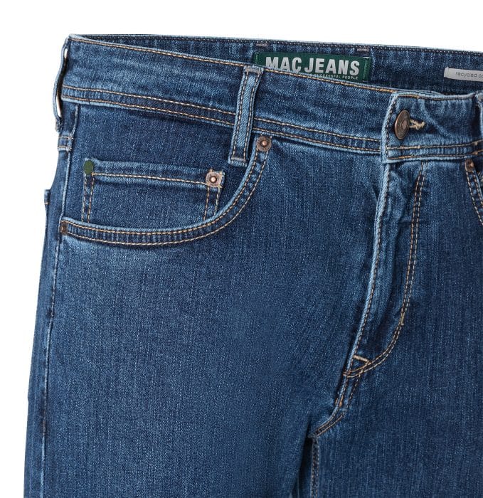 Close-up of blue denim jeans with visible stitching, front button, and pockets. Label inside waistband reads MAC JEANS in green and white text. The jeans show a classic five-pocket design.