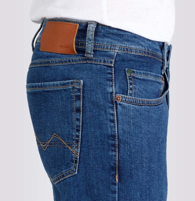 A close-up side view of a person wearing blue denim jeans and a white shirt. The jeans have a brown leather patch on the waistband, visible stitching, and back and front pockets.