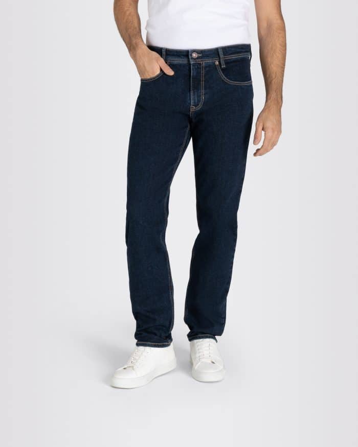 A man wearing a white T-shirt, dark blue jeans, and white trainers stands with one hand in his pocket against a plain light grey background.