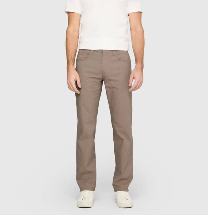 A person wearing a white short-sleeve shirt, light brown straight-leg trousers, and white trainers stands against a plain light grey background.