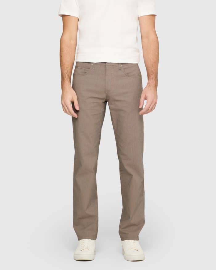 A person wearing a white short-sleeve shirt, light brown straight-leg trousers, and white trainers stands against a plain light grey background.