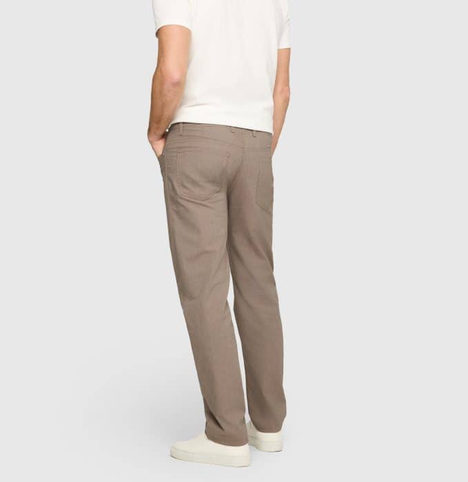 A person wearing a white short-sleeve shirt, light brown trousers, and white trainers stands facing away with hands in their back pockets against a plain light grey background.