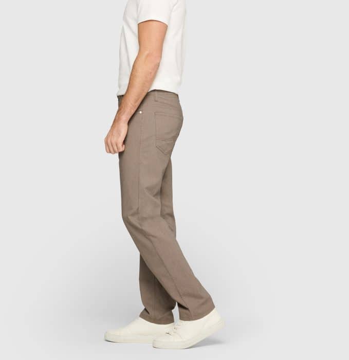 A person stands sideways wearing a white T-shirt, light brown trousers, and white trainers against a plain light grey background. The person's head is not visible.