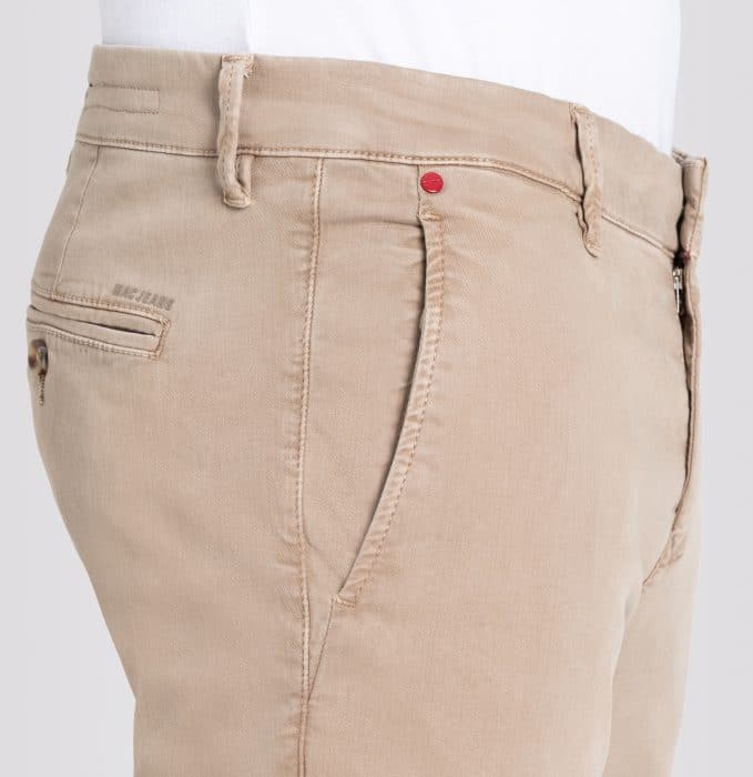 Close-up of a person wearing light beige chino trousers with side and back pockets, a visible waistband, belt loops, and a small red rivet near the pocket. A white shirt is tucked in at the waist.