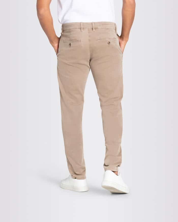 A person wearing tan chinos, a white shirt, and white trainers stands facing away with hands in pockets, against a plain light background.