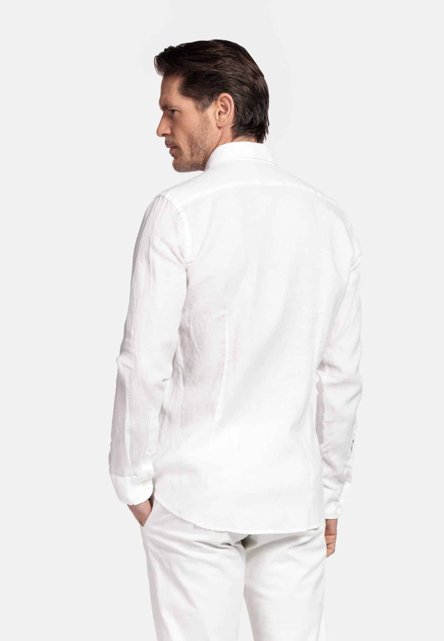 A man with short brown hair is standing and facing away from the camera, wearing a long-sleeved white button-up shirt and white trousers against a plain light background.