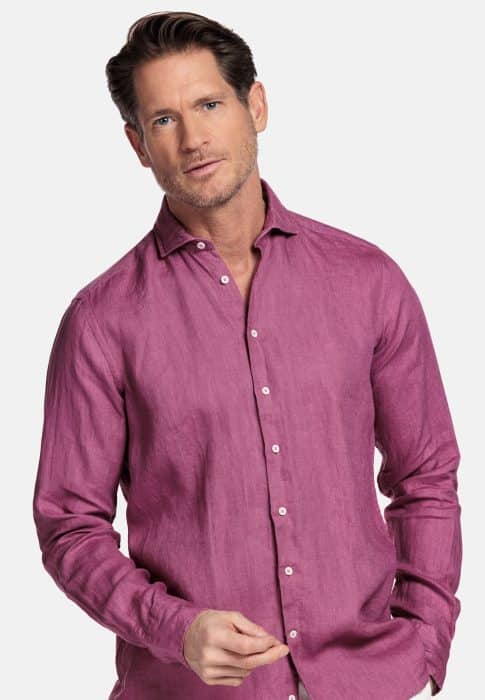 A man with short brown hair and light stubble wears a long-sleeved, button-up magenta shirt. He stands against a plain white background, looking forward with a neutral expression.