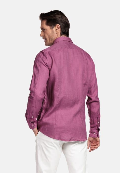 A man facing away, wearing a long-sleeved, buttoned purple shirt and white trousers, standing against a plain light background.