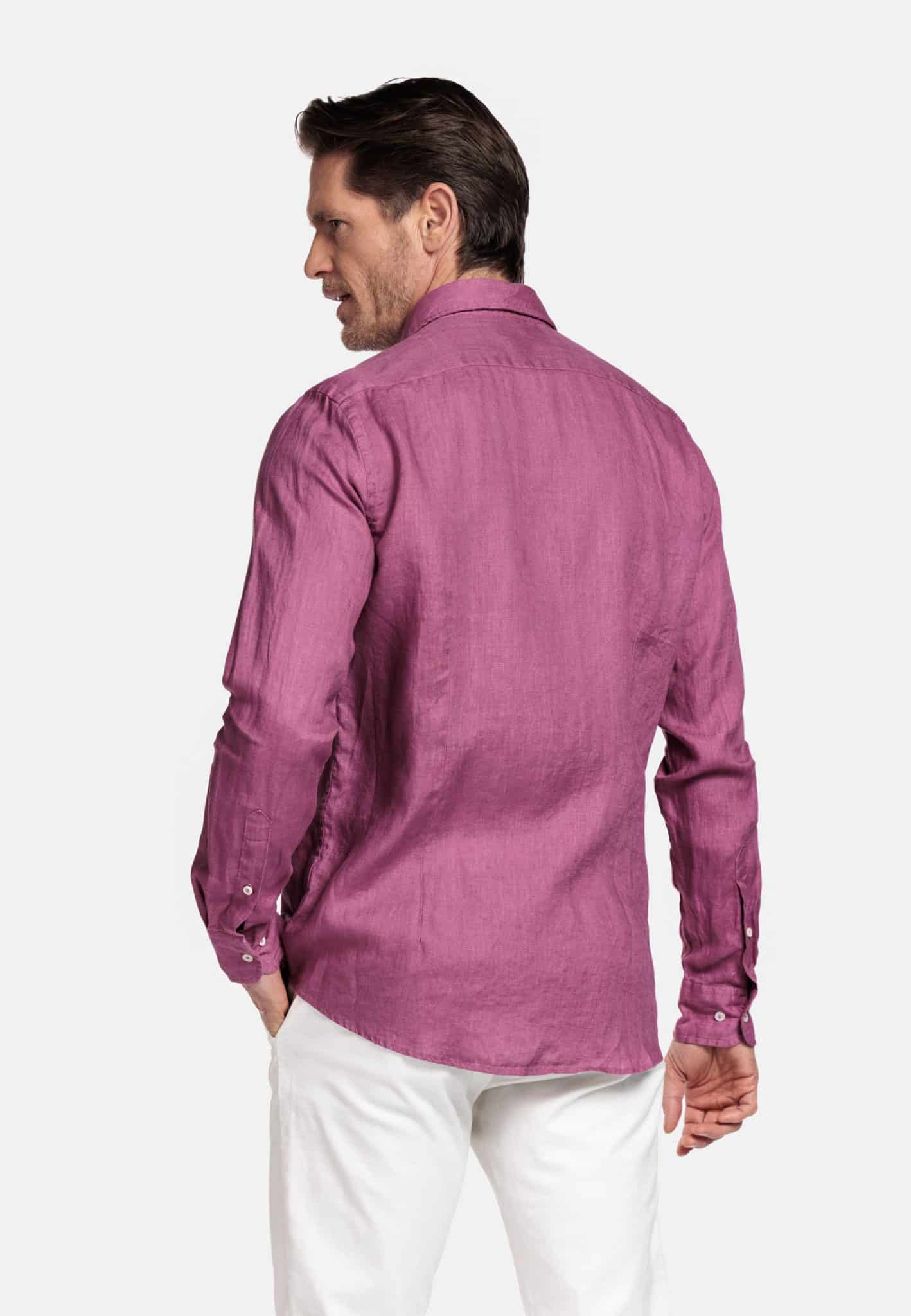 A man facing away, wearing a long-sleeved, buttoned purple shirt and white trousers, standing against a plain light background.
