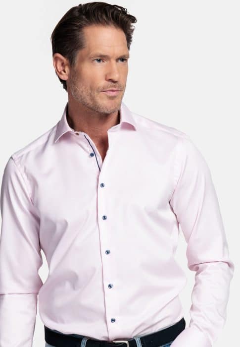 A man wearing a light pink dress shirt with dark buttons, dark jeans, and a black belt stands against a plain white background, looking slightly to his right.