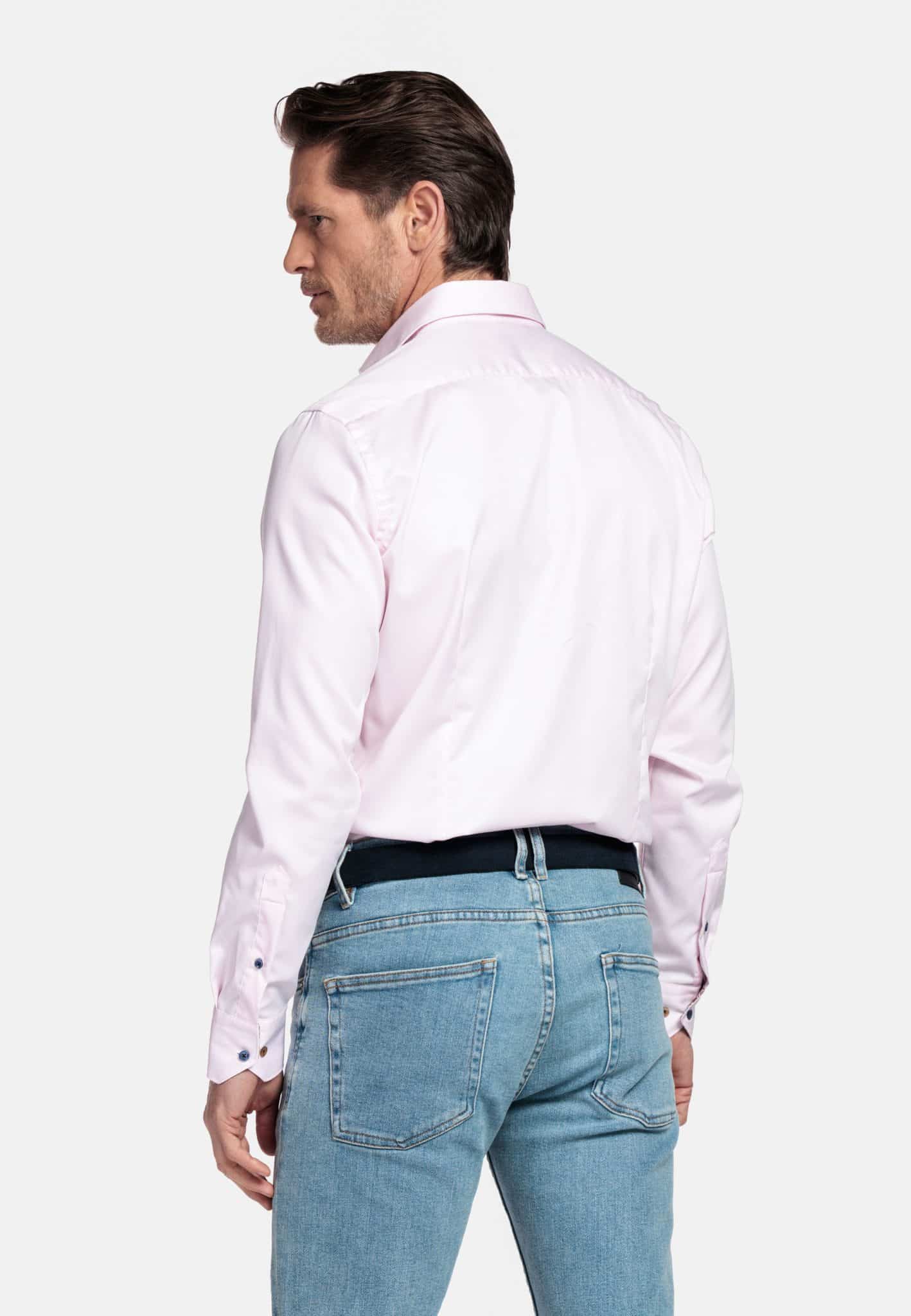 A man stands with his back to the camera, wearing a light pink long-sleeved dress shirt and light blue jeans, against a plain white background.
