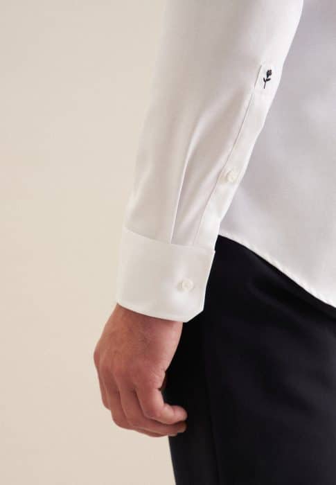 A person wearing a white long-sleeved dress shirt and black trousers, with a focus on the left arm and shirt cuff, which shows a small embroidered detail near the button.