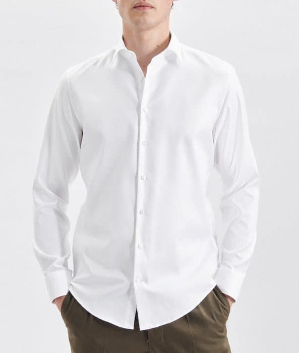 A person wearing a long-sleeved, white button-up shirt and brown trousers stands with hands in their pockets against a plain white background. Their face is partially out of frame.