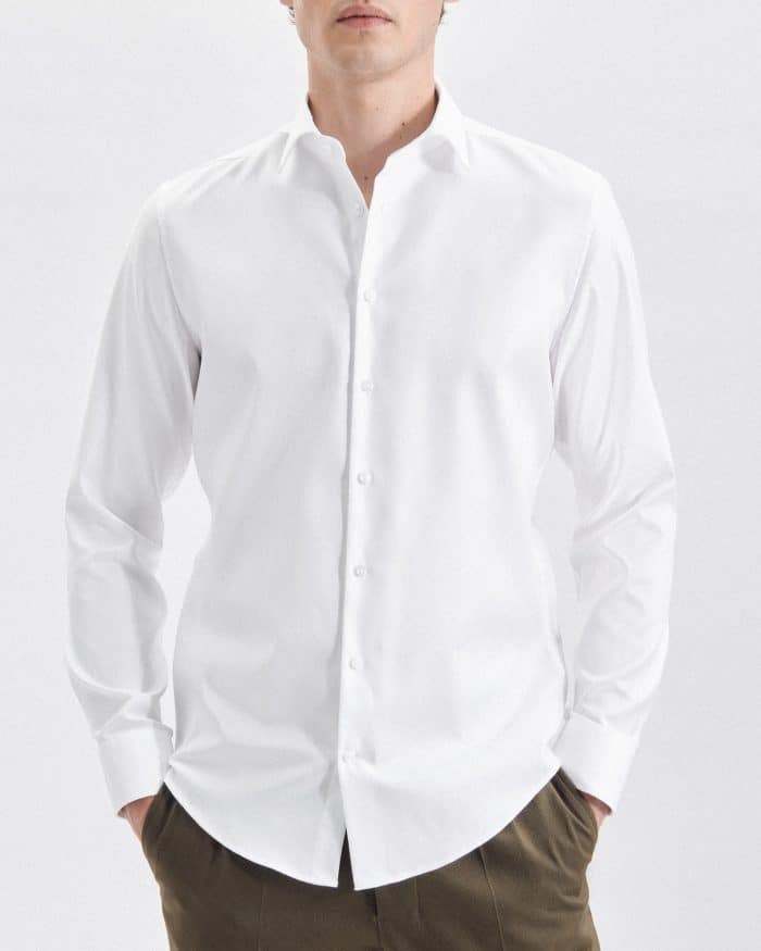A person wearing a long-sleeved, white button-up shirt and brown trousers stands with hands in their pockets against a plain white background. Their face is partially out of frame.