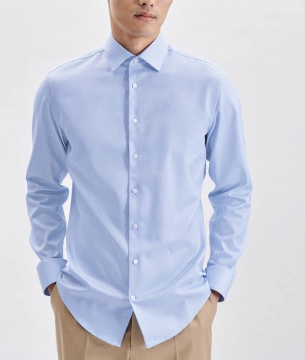 A man wearing a light blue dress shirt with white buttons and beige trousers stands with his hands in his pockets against a plain, light background.