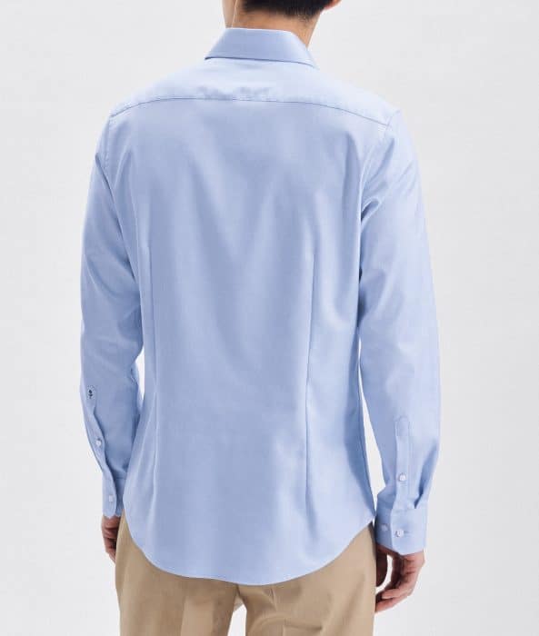 A man is shown from the back wearing a light blue long-sleeved button-up shirt and beige trousers, standing against a plain white background.