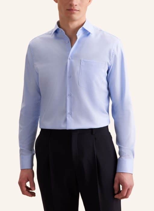 A man wearing a light blue long-sleeve dress shirt with a chest pocket and black dress trousers, standing against a plain light background. His face is partially out of frame.