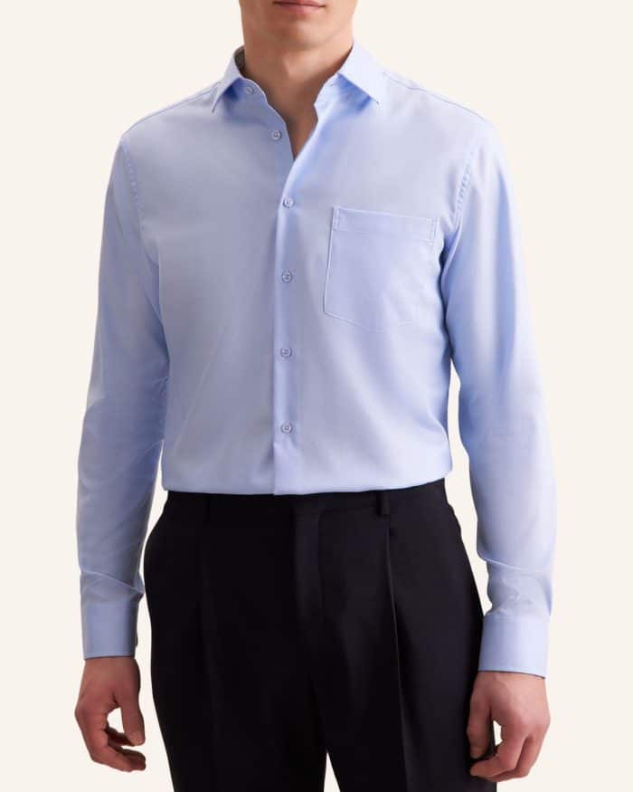 A man wearing a light blue long-sleeve dress shirt with a chest pocket and black dress trousers, standing against a plain light background. His face is partially out of frame.