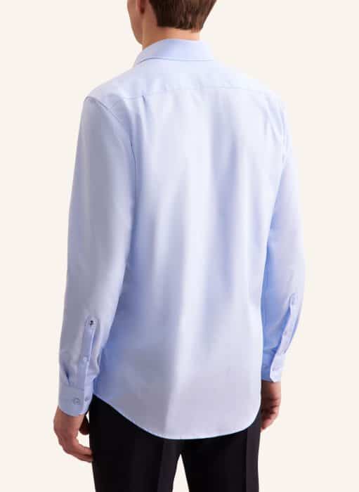 A person wearing a light blue, long-sleeved dress shirt and dark trousers is shown from the back against a plain background. The shirt is untucked and features buttoned cuffs.