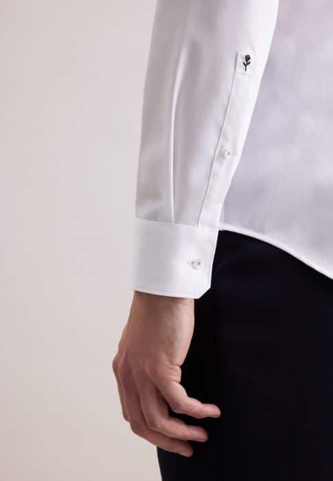 A person wearing a white dress shirt with buttoned cuffs and black trousers. Only the lower arm, cuff, and hand are visible. The shirt cuff has a small floral embroidery detail near the button.