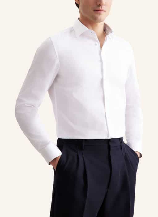 A man wearing a crisp white long-sleeved dress shirt and dark trousers stands with his hands in his pockets, facing slightly to the side against a plain background.