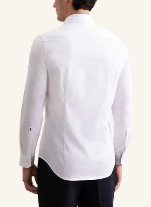 A man is shown from the back wearing a long-sleeved, white dress shirt with a collar and black trousers. The shirt has buttoned cuffs and a curved hem. The background is plain and light-coloured.