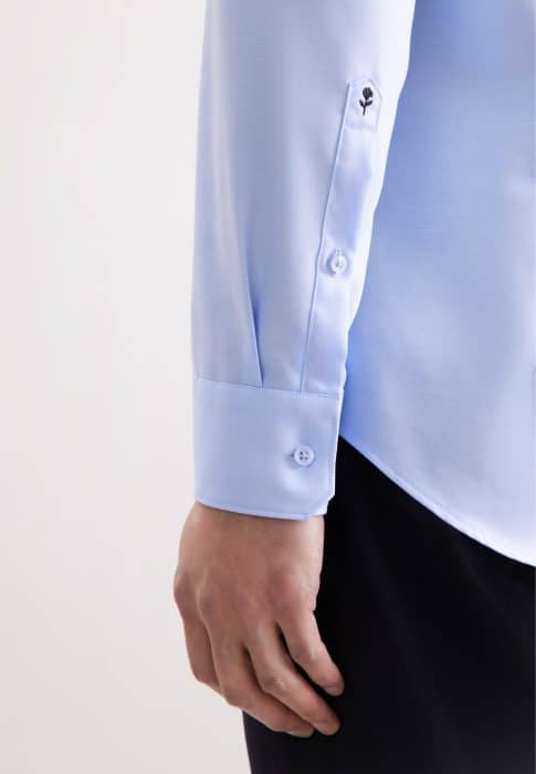 A person wearing a light blue long-sleeve dress shirt with buttoned cuffs and dark trousers. The image focuses on the lower sleeve and hand against a plain background.