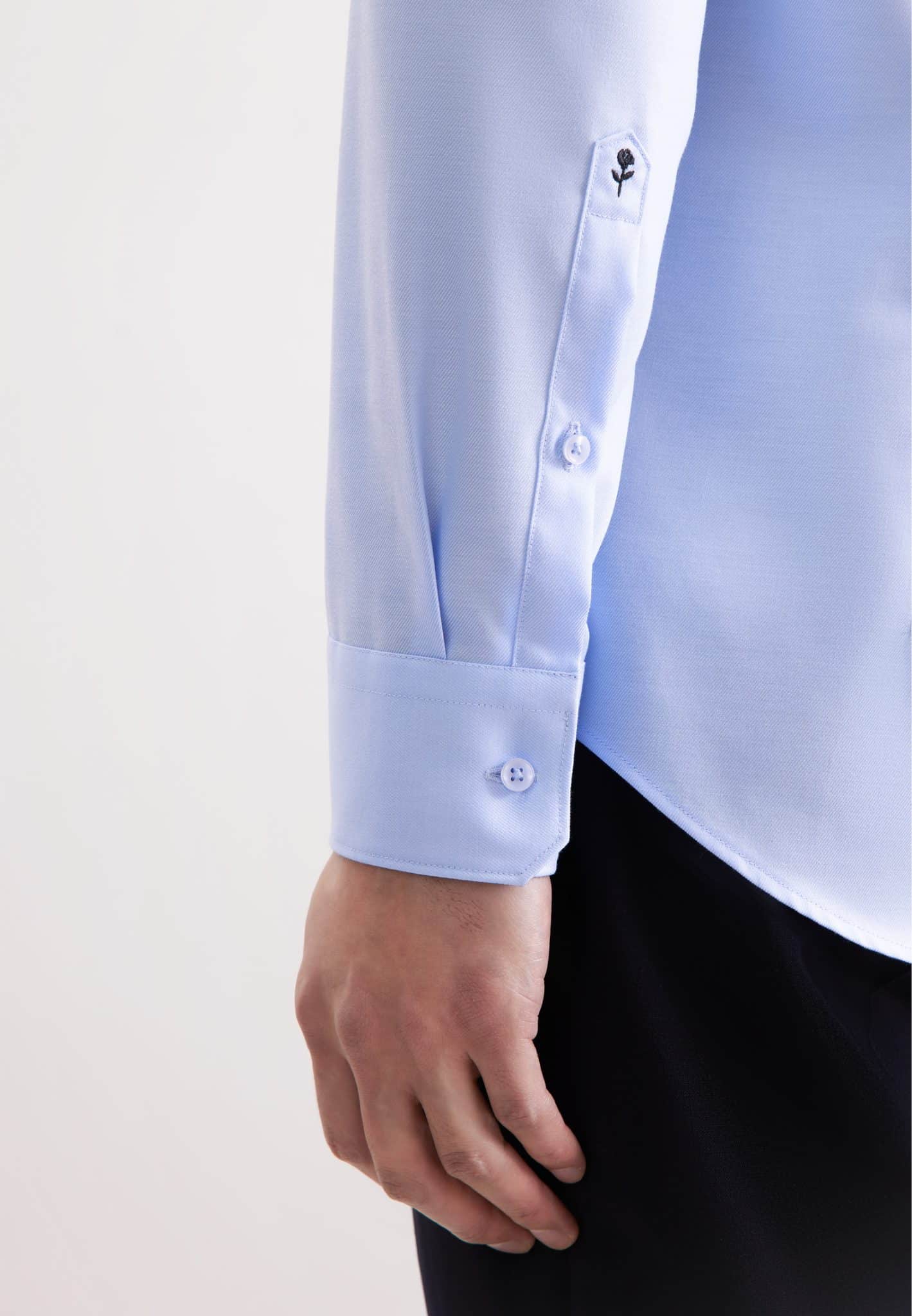 A person wearing a light blue long-sleeve dress shirt with buttoned cuffs and dark trousers. The image focuses on the lower sleeve and hand against a plain background.