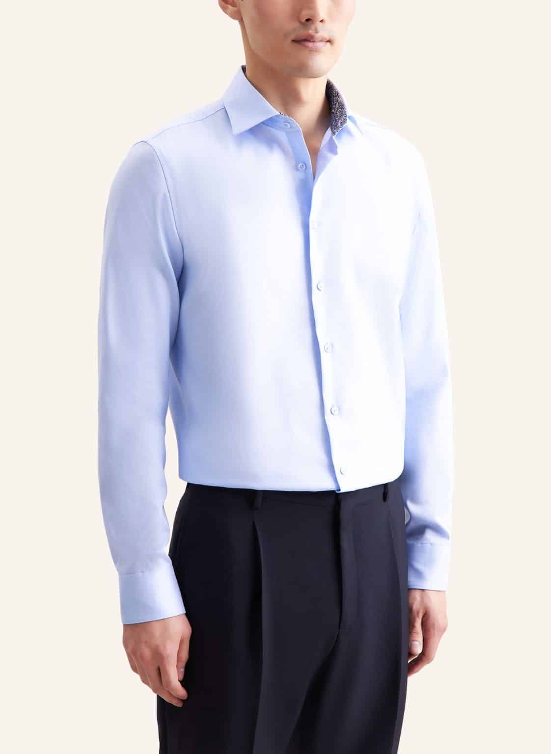 A man wearing a light blue, long-sleeved dress shirt tucked into black dress trousers, standing against a plain light background. His face is partially out of frame and he has a relaxed posture.
