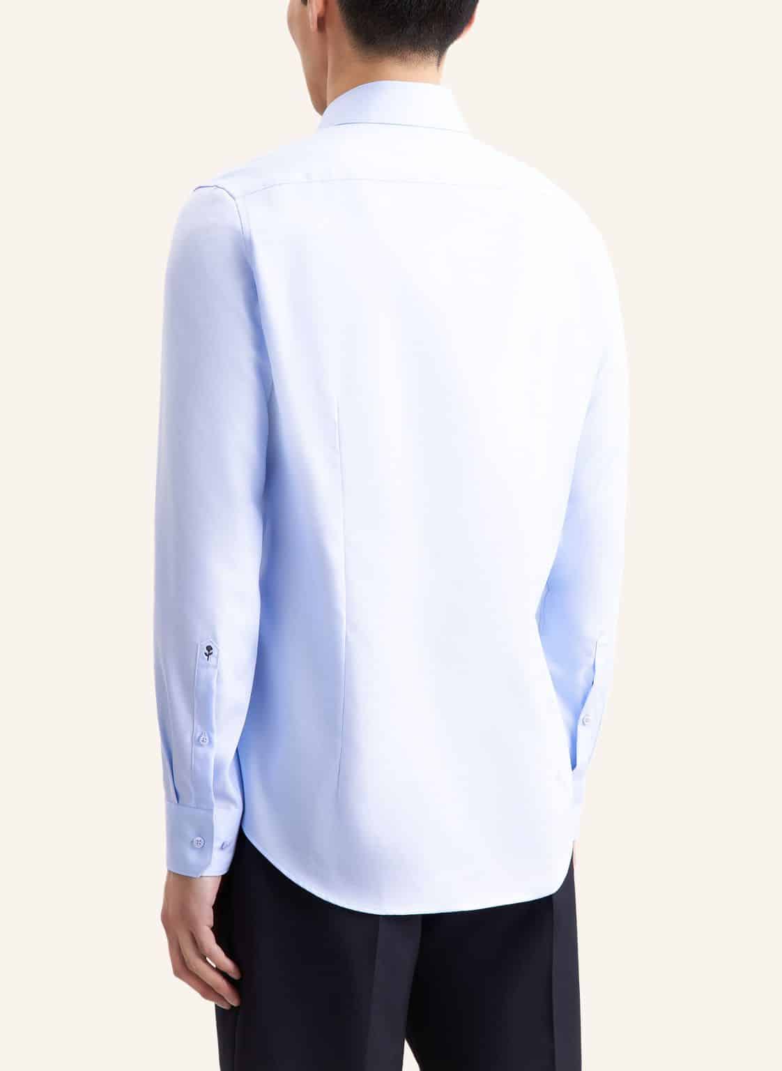 A person is shown from the back wearing a light blue long-sleeved dress shirt and black trousers. The shirt has buttoned cuffs and a smooth, tailored fit. The background is plain and light-coloured.