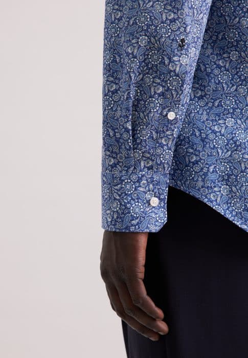 A close-up of a person's arm wearing a long-sleeved blue paisley-patterned shirt with white buttons, shown against a plain light background. The person's hand is relaxed and visible near dark trousers.