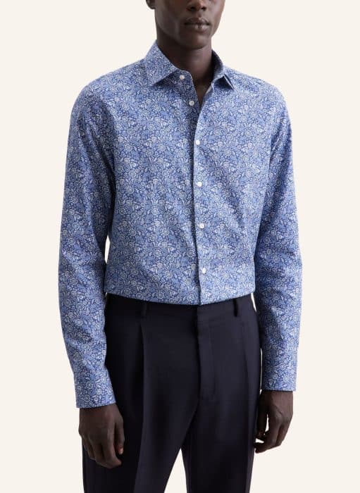 A man wearing a blue long-sleeved button-up shirt with a small floral pattern and black trousers, standing against a plain light background.