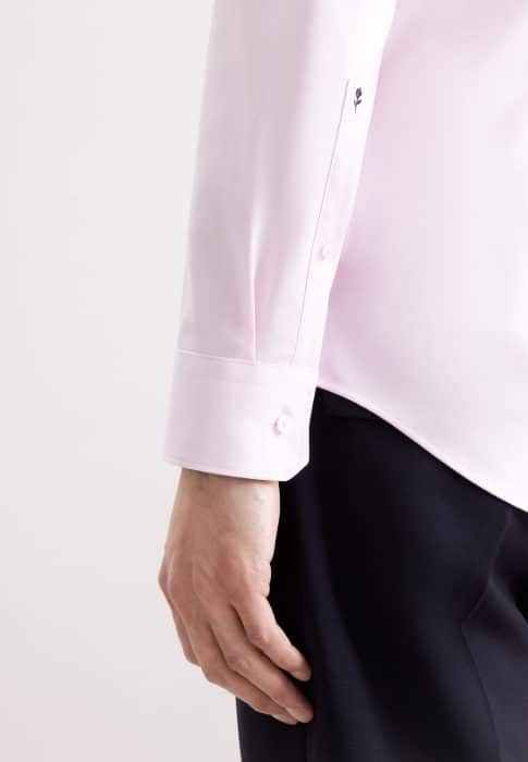 A person wearing a light pink long-sleeved dress shirt with buttoned cuffs and dark trousers, shown from the waist down with their arm relaxed at their side.