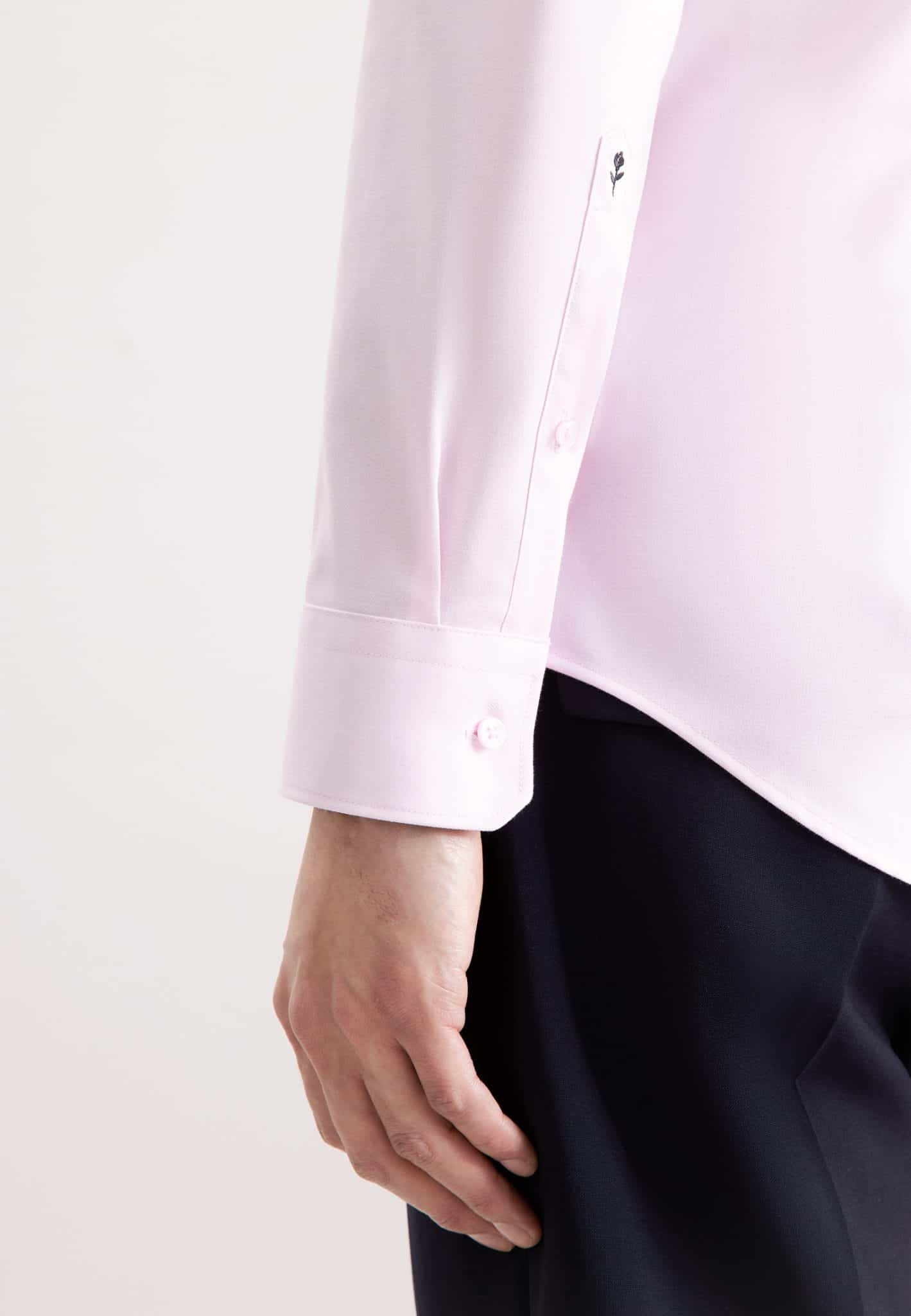 A person wearing a light pink long-sleeved dress shirt with buttoned cuffs and dark trousers, shown from the waist down with their arm relaxed at their side.