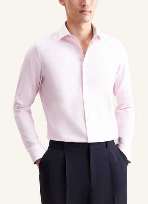 A person wearing a light pink button-up dress shirt with long sleeves and dark pleated trousers, standing with hands in their pockets against a plain light background.