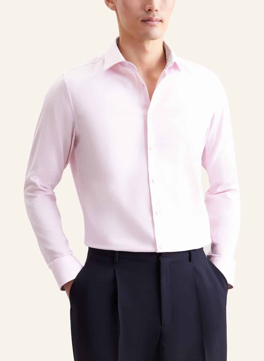 A person wearing a light pink button-up dress shirt with long sleeves and dark pleated trousers, standing with hands in their pockets against a plain light background.