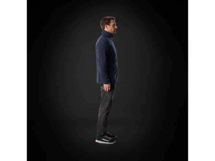 A man stands in profile against a dark background, wearing a navy jacket, dark jeans, and black trainers with white soles.
