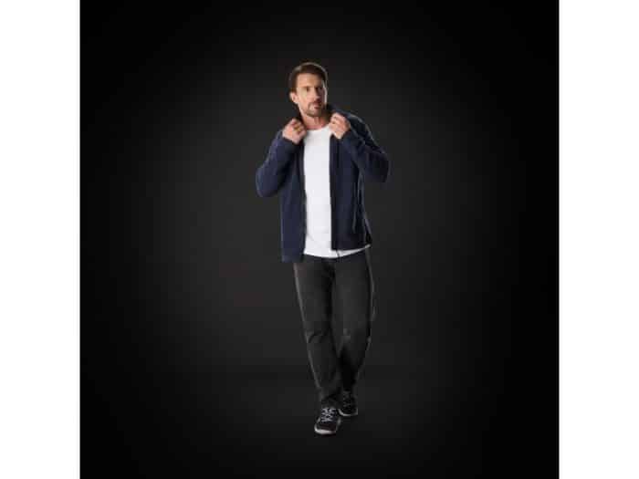 A man stands against a dark background, wearing a navy jacket over a white shirt, black trousers, and black trainers. He holds the jacket collar with both hands and looks slightly to the side.