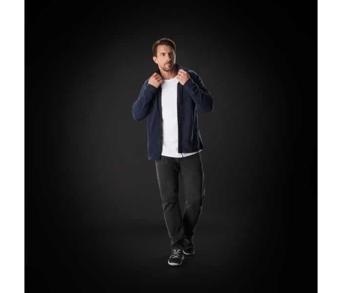 A man stands against a dark background, wearing a navy jacket over a white shirt, black trousers, and black trainers. He holds the jacket collar with both hands and looks slightly to the side.