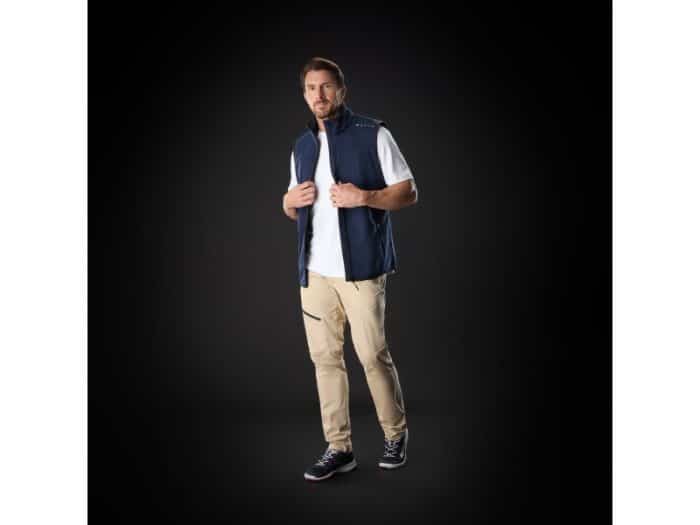 A man stands against a black background, wearing a navy blue waistcoat over a white T-shirt, beige trousers with a zippered pocket, and black trainers with white soles. He is looking forward and holding his waistcoat collar.