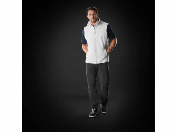 A man stands against a dark background, wearing a white zip-up gilet over a navy shirt, dark trousers, and black trainers. His hands are in his gilet pockets, and he looks slightly to the side with a neutral expression.