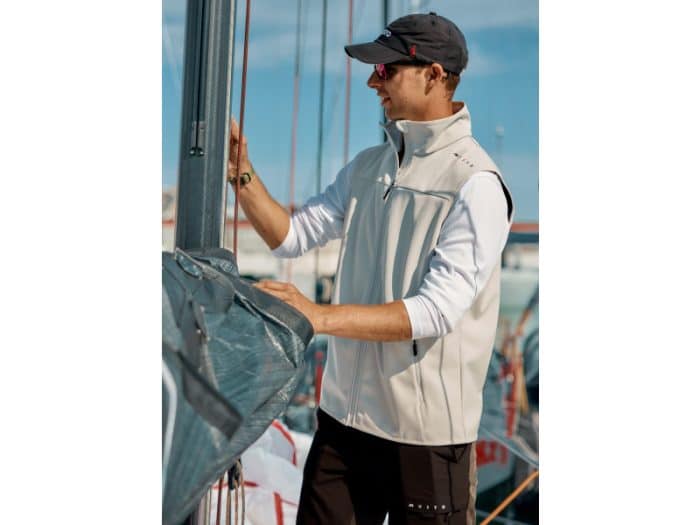 A man wearing sunglasses, a cap, and a light-coloured waistcoat stands on a sailboat, adjusting or inspecting equipment with a marina and other boats visible in the background.