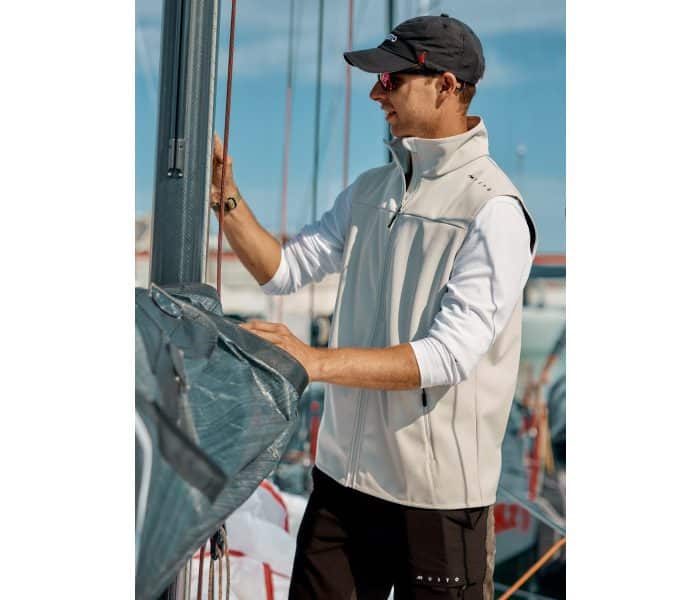 A man wearing sunglasses, a cap, and a light-coloured waistcoat stands on a sailboat, adjusting or inspecting equipment with a marina and other boats visible in the background.