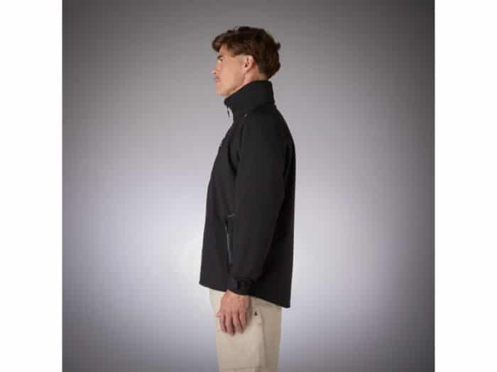 A man is standing in profile, facing left, wearing a black jacket with a high collar and light-coloured trousers against a plain, gradient grey background.